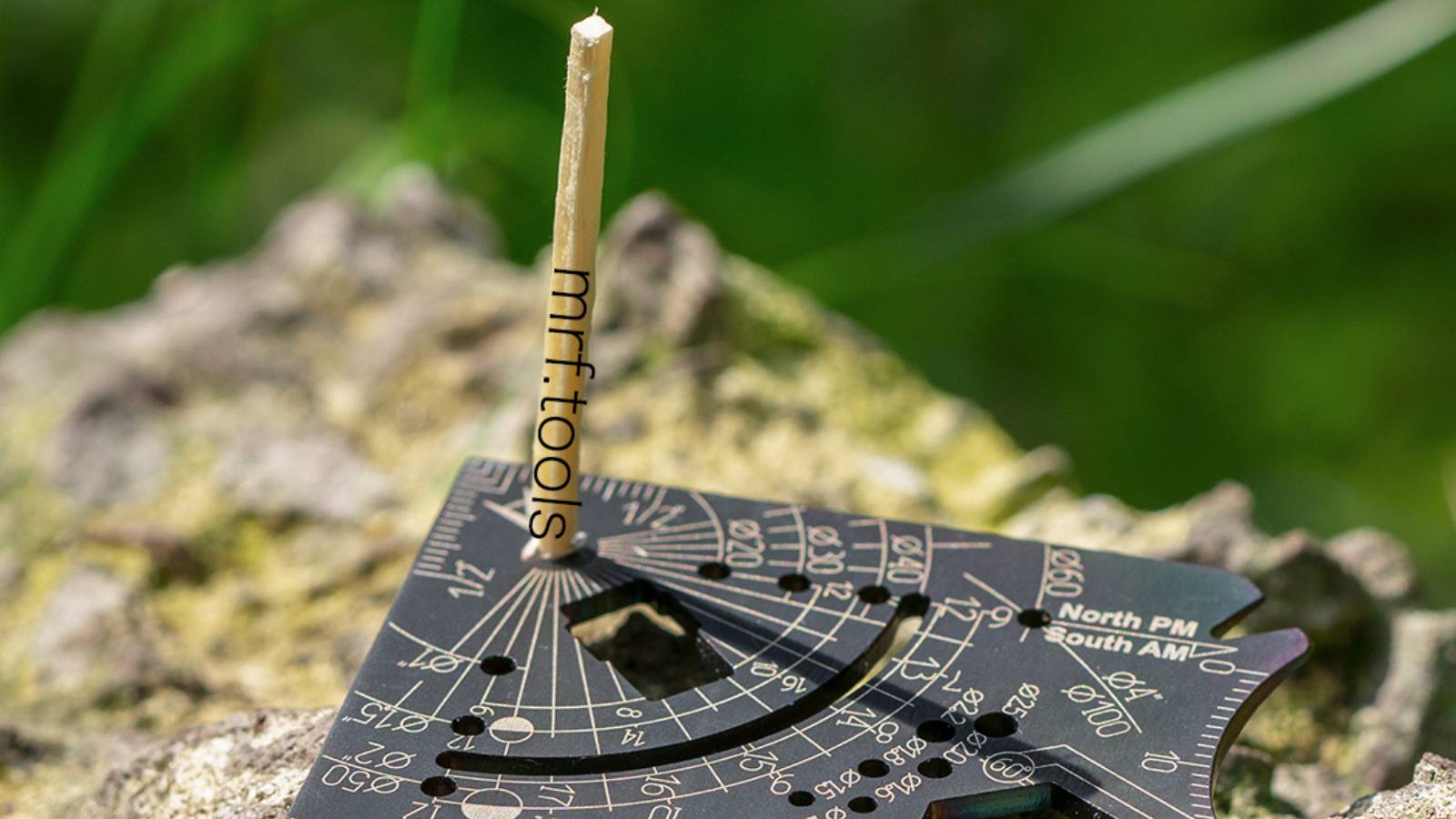 Black card resting on a mossy rock in sunlight with a matchstick standing in its center hole, the shadow falling across the engraved sundial scale